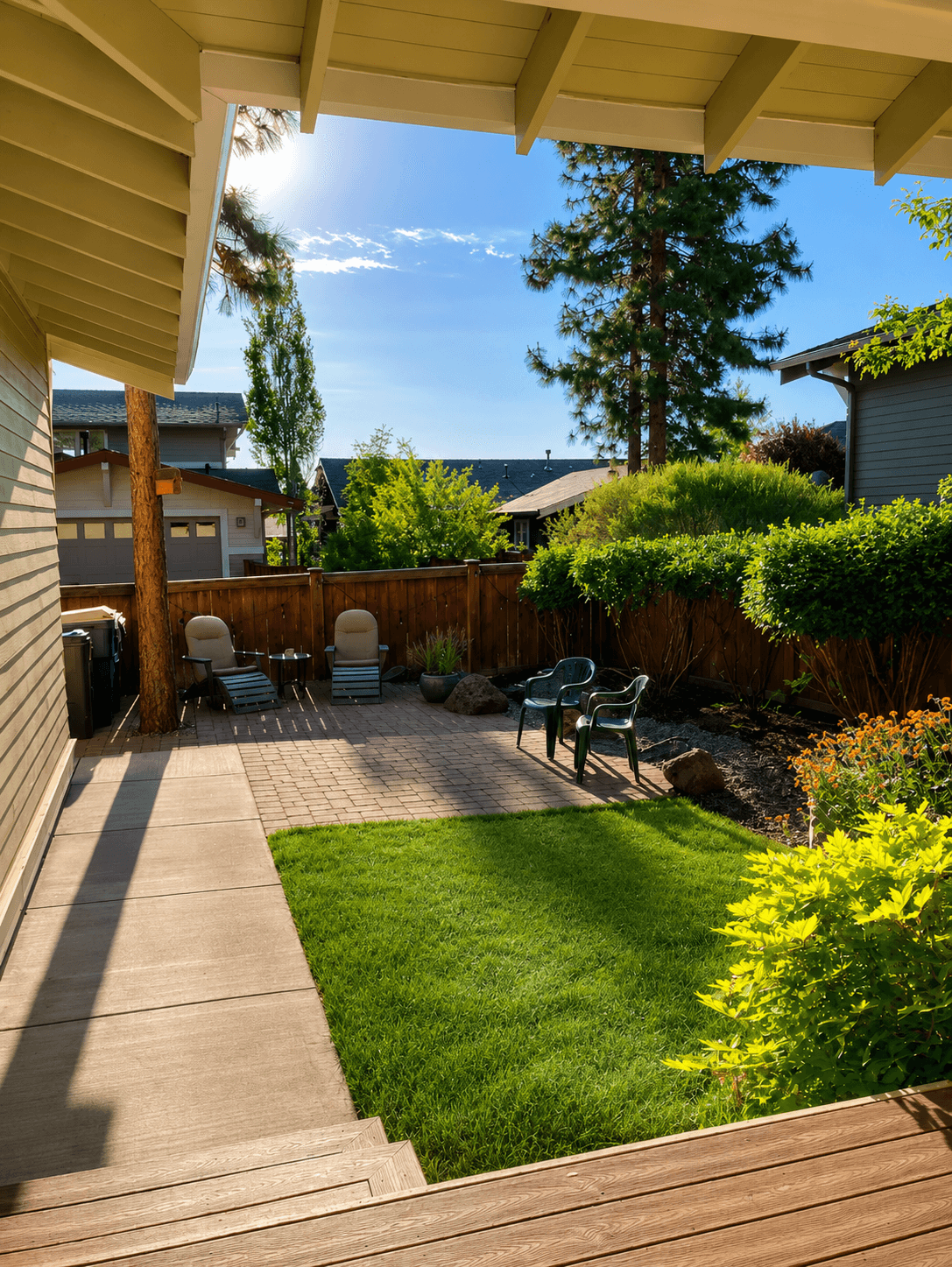 Sunny backyard featuring a brick patio, green lawn, outdoor seating, and tall pine trees.