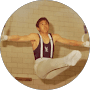 Male gymnast in a dark singlet performing an L-sit on rings against a brick wall.